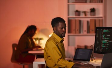 Portrait of African-American IT developer writing code on multiple computer screens while working in office, copy space